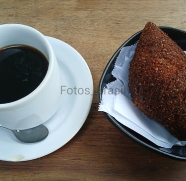 Uma xícara de café fumegante sobre um pires branco, ao lado num pratinho, um kibe dourado e crocante
