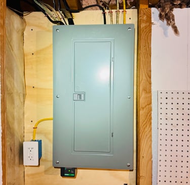 A modern grey residential electrical panel board installed on plywood with visible wiring and a surge protector.