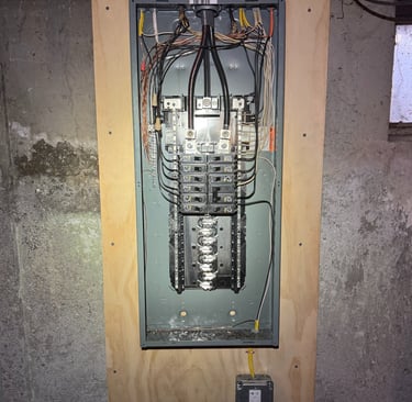 Open residential electrical panel with circuit breakers and wiring mounted on a plywood board.