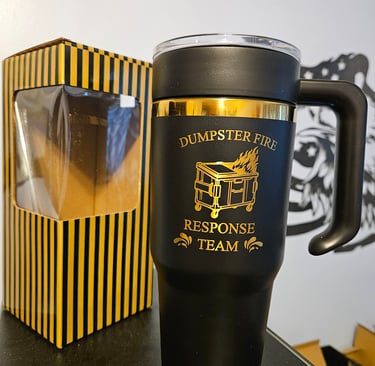 Black and gold Dumpster Fire Response Team insulated travel mug with handle next to a yellow striped gift box.