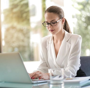 a woman in a white shirt and glasses is sitting at a table and working on laptop