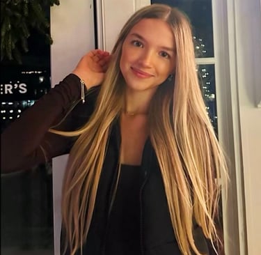 Emily smiling warmly indoors at night her long blonde hair as lights twinkle in the background
