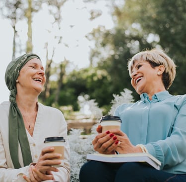 two women sitting on a bench in a park laughing and drinking coffee