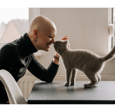 a smiling woman in a black turtle neck and a  bald head and a grey cat are touching nose to nose