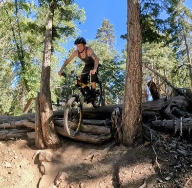 Mark Hower riding a mountain bike over a fallen log in a pine forest in Southern California, black