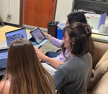 Three students sitting at a table, each with a computer or tablet in front of them reviewing video