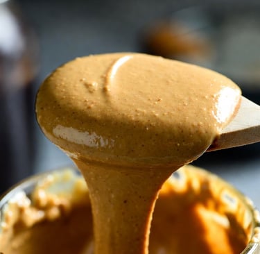 Primer plano de una cuchara de madera goteando mantequilla de almendras cremosa sobre un tarro de cristal - Raw Rico
