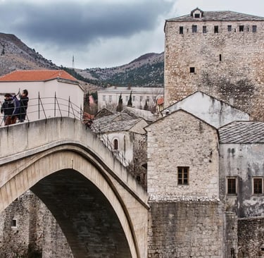 Tourists crossing Stari Most stone bridge in Mostar with mountains and cloudy sky
