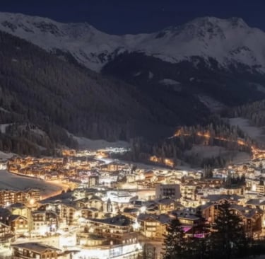 A panoramic, nighttime view of an Alpine ski resort town nestled in a valley, with glowing lights vi