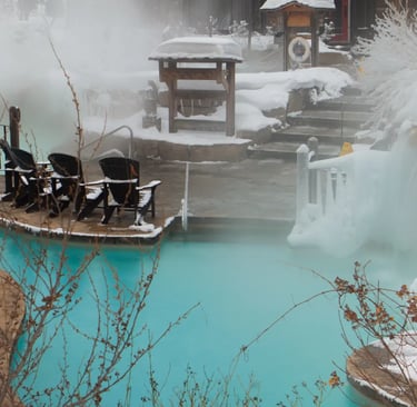 A large outdoor thermal spa complex with steaming pools surrounded by snow-covered mountains and pin