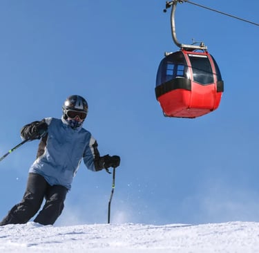 A skier carves down a bright, snowy slope while a vibrant red gondola passes overhead against a clea