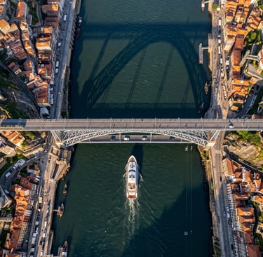 Aerial view of luxury yacht passing under historic Douro River bridge in Porto at golden hour.
