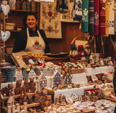 A smiling vendor at a Christmas market stall piled high with various handmade goods and small souven