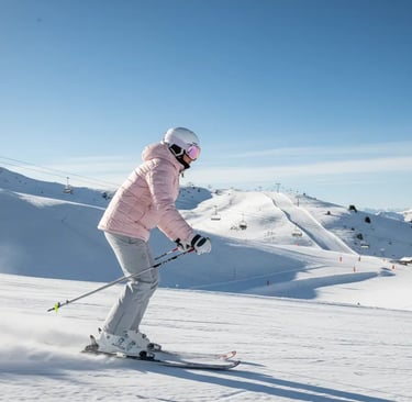 A woman skiing down a snowy slope in a light pink puffer jacket and grey ski pants, showcasing a fem