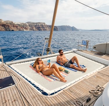 Couple sunbathing on catamaran bow netting with Greek island cliffs in background