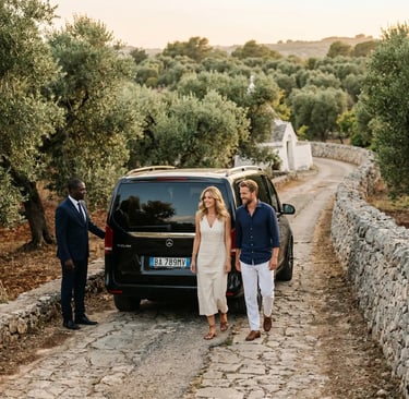 Chauffeur and couple beside black Mercedes V-Class on stone road through Puglian olive groves.