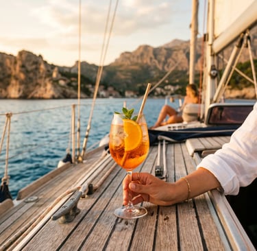 Hand holding Aperol Spritz glass on sailboat deck with rocky coastline at golden hour