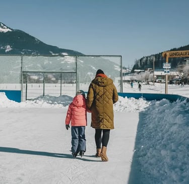 A parent and a young child in a pink jacket holding hands while walking onto a sunny outdoor ice ska