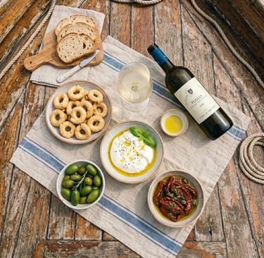 Overhead flat-lay of burrata, taralli, olives, sun-dried tomatoes and Puglia white wine on boat deck