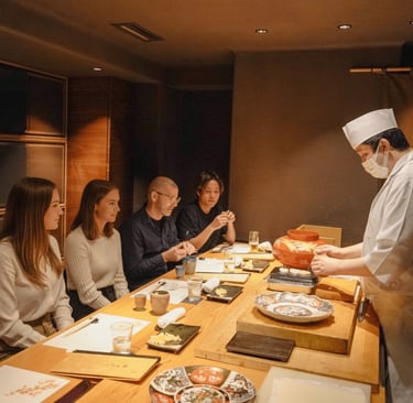 A chef in a white uniform and face mask serves food to four seated people at a wooden table with dis