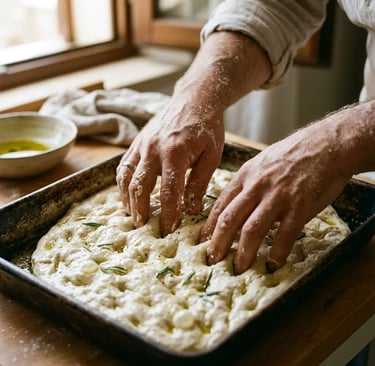 Hands pressing dimples into focaccia dough with olive oil and rosemary in an iron tin
