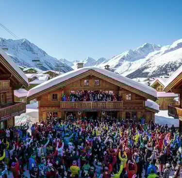 Après-ski party at a mountain chalet with a large crowd and scenic snowy peaks in the background.