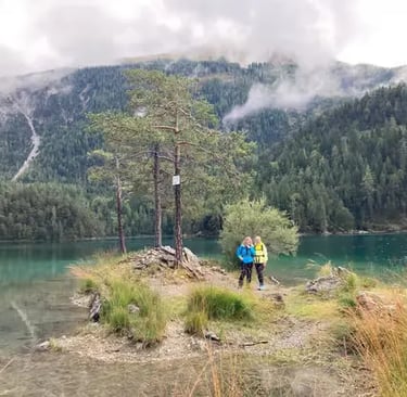 Two hikers standing near turquoise alpine lake with pine trees and misty mountain peaks behind