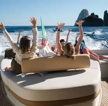 Guests with arms raised on yacht aft deck approaching Faraglioni rocks off Capri