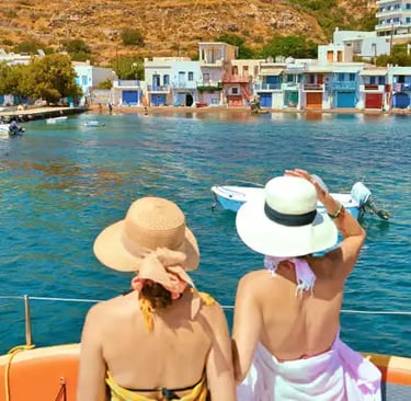 Two women in sun hats on catamaran approaching colorful Klima fishing village in Milos