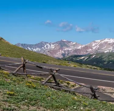 Winding asphalt road with wooden fences overlooking snow-capped Rocky Mountains.
