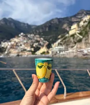 Hand holding traditional lemon ceramic cup on boat with Positano village and mountains in background