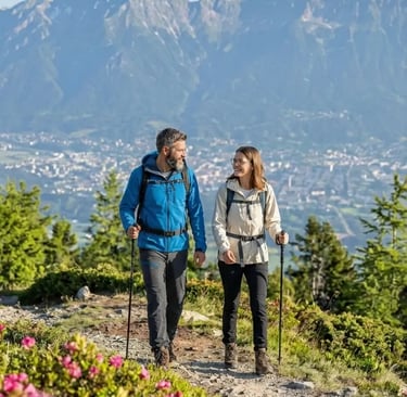 Hikers walk past vibrant pink wildflowers with the Austrian Alps spanning the horizon.