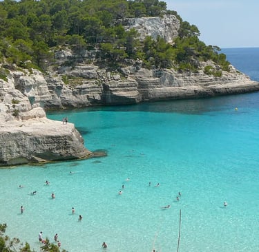 Swimmers in crystal-clear turquoise waters of a pine-fringed cove in Menorca, Spain