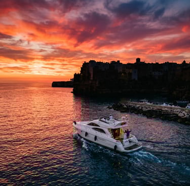 White private yacht leaving Polignano a Mare harbor under dramatic red sunset sky