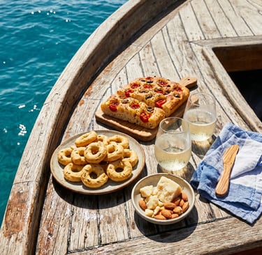 Taralli, focaccia barese, cheese, almonds and prosecco on wooden boat deck over turquoise sea.