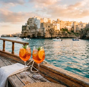 Two Aperol spritz glasses on boat deck with Polignano a Mare at sunset
