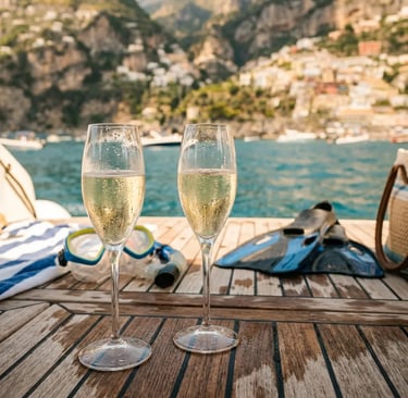 Two Prosecco glasses on teak boat deck with snorkeling gear and Positano coastline behind