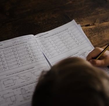 a child's hand holding a pencil and writing on a Maths work book