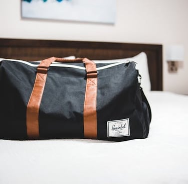 Black Herschel duffel bag with brown leather straps sitting on a white hotel bed.