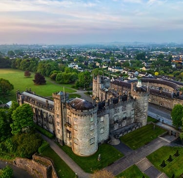 Aerial view of Kilkenny Castle in Ireland surrounded by lush green gardens and a historic city skyline.
