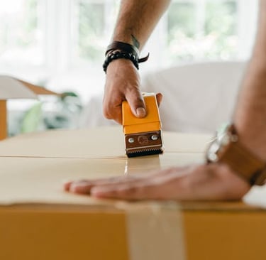 A person using a yellow tape dispenser to seal a cardboard moving box for shipping or relocation.