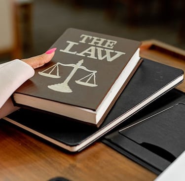 A person holding a brown book titled The Law with scales of justice on a courtroom desk.