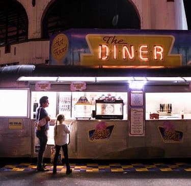 Customers ordering food from a vintage silver Airstream food truck diner with neon signs at night.