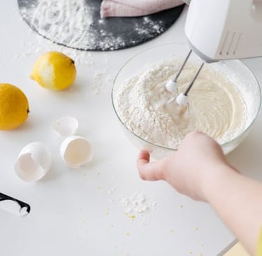 A person using an electric hand mixer to blend cake batter in a glass bowl with fresh lemons and eggshells.