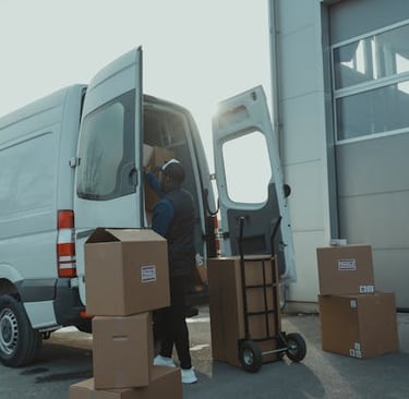 A delivery driver loading cardboard shipping boxes into a white cargo van outside a warehouse.