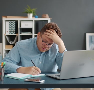 a man sitting at a desk with a laptop and a notebook, thinking how he can save money as a student
