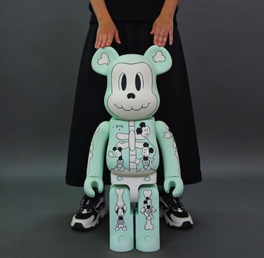 A mint green Bearbrick 1000% figure with Mickey Mouse skeleton graphics being held by a person.