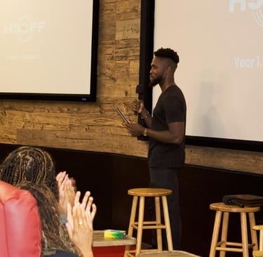 Tim Salau and Krishna Lyles stand in front of an audience with a stool in front of them.