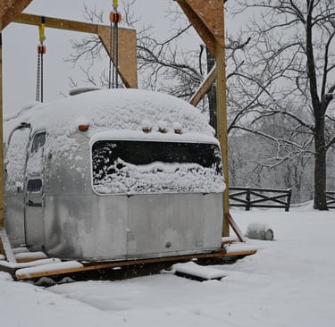 Snowcovered shell of the airstream sitting on the lumber. 