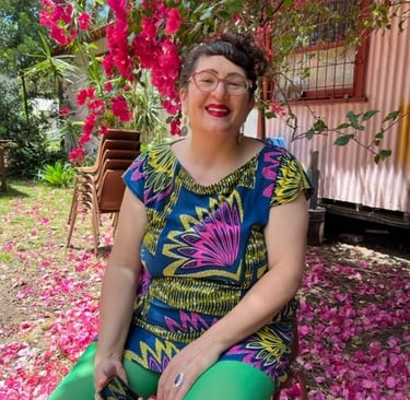 A person with curly brown hair and light skin sits under a tree smiling.